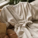 Robe en lin femme écru près d’une fenêtre, tissu froissé et ceinture nouée